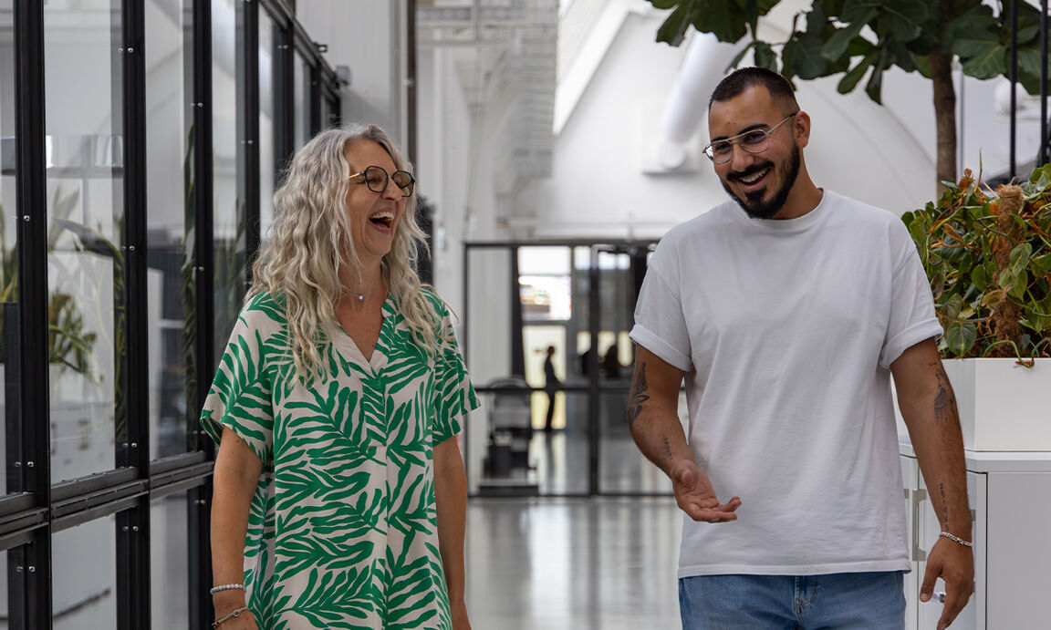 Two colleagues, a woman and a man, walking down a hallway in a modern open-plan office.