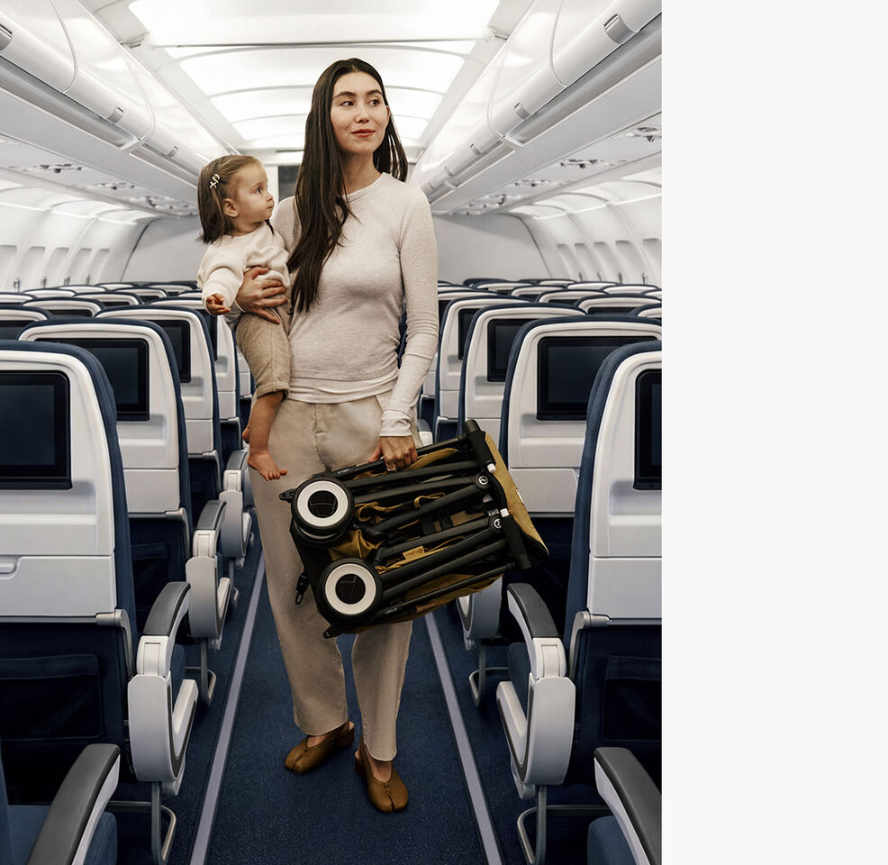 A mother on a plane amidst plane seats, holding her daughter with one hand and a folded Libelle with the other.