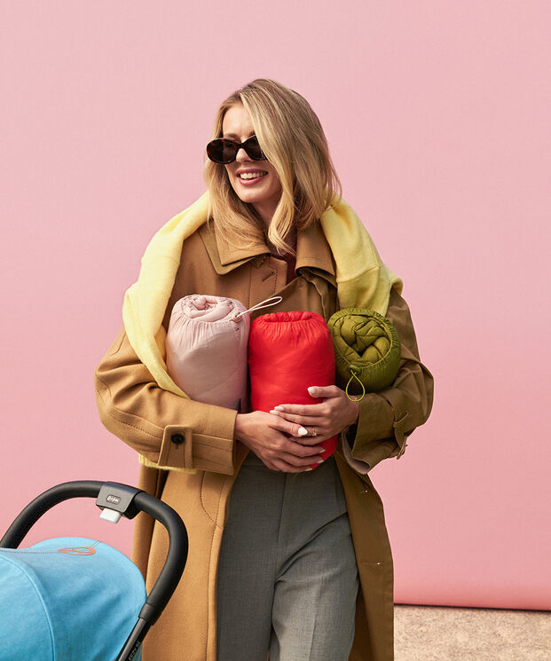 A woman wearing sunglasses holds three colourful blankets in her arms, against a pink background.